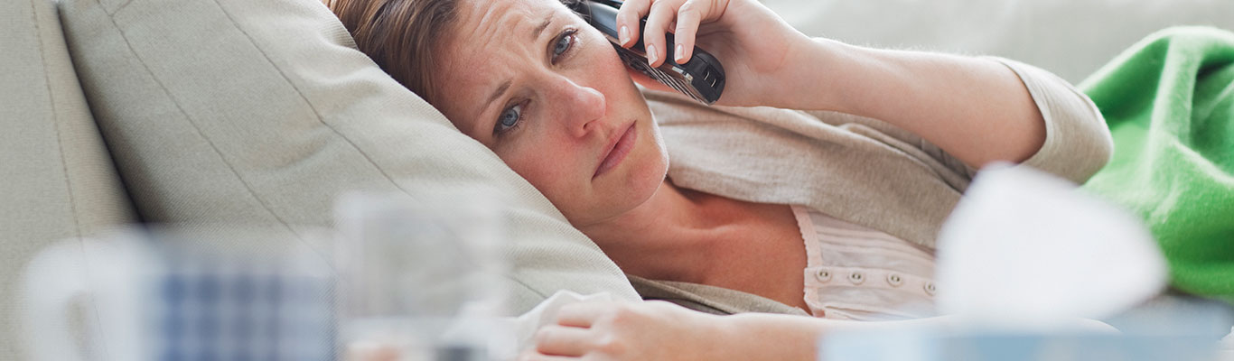 Woman lying on the couch with a box of tissue and mug while making a phone call.