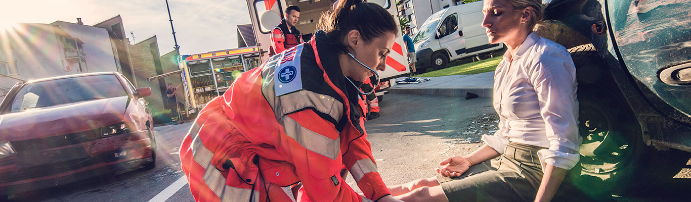 Disheveled woman sitting on the road, leaning against a car wheel well with a first responder attending to her injuries after what appears to be a car accident.