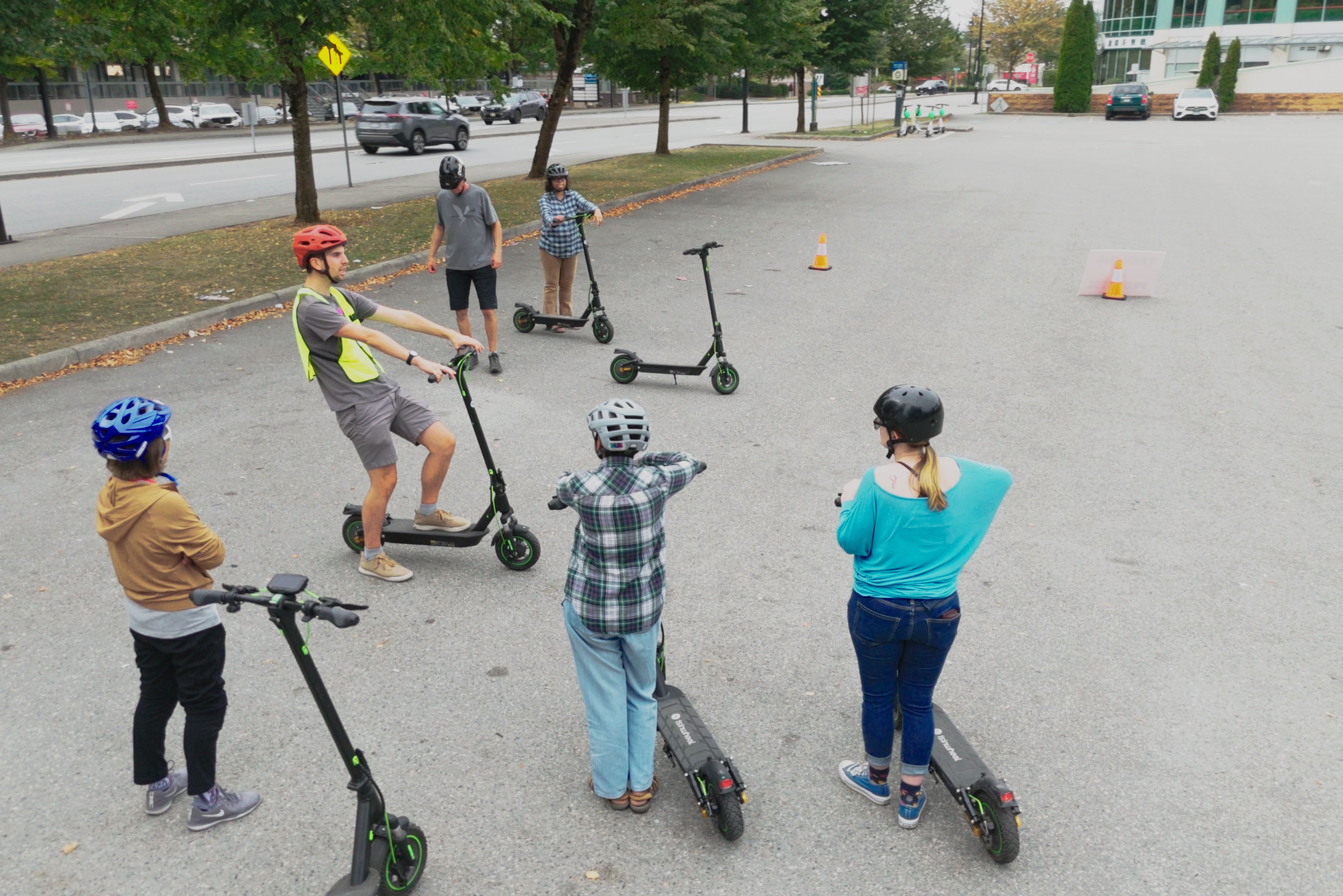 A group of teens with e-scooters gather around an instructor for an e-scooter lesson in a parking lot