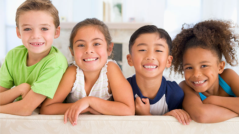 Photo of four smiling children leaning over the back of a sofa - two boys and two girls.
