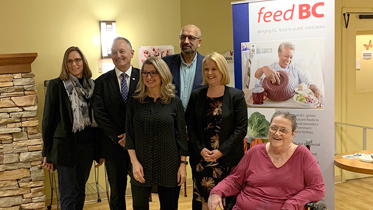 Group of diverse adult women and men standing with elder woman seated in wheelchair in front of Feed BC sign