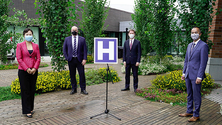 BC Cancer Centre announcement at the new Surrey Hospital. A sign with the letter "H" in the center of the picture. Dr. Victoria Lee and Premier John Horgan wearing masks standing to the left of the sign. Minister Adrian Dix and Dr. Kim Nguyen Chi wearing masks standing to the right of the sign.