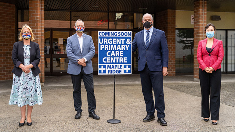 A sign for the new urgent and primary care centre opening soon in Maple Ridge in the center of the picture. Lisa Beare, MLA and Bob D'Eith MLA wearing masks standing to the left of the sign. Premier John Horgan and Dr. Victoria Lee wearing masks standing to the right of the sign. 