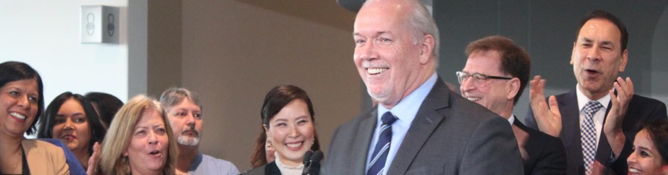 Premier John Horgan smiling at the announcement for the New Surrey Hospital with Dr. Victoria Lee and Minister Adrian Dix and others in the background